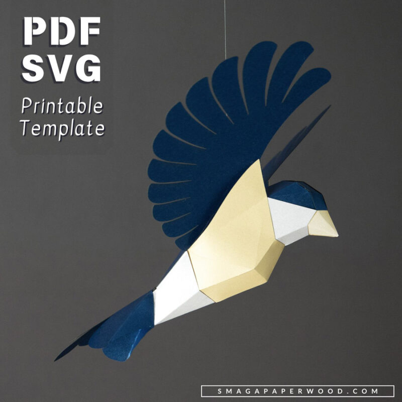 Geometric papercraft bird sculpture suspended in mid-flight against a dark gray background. The bird is crafted from blue, white, and yellow paper, featuring layered wings and a faceted body.