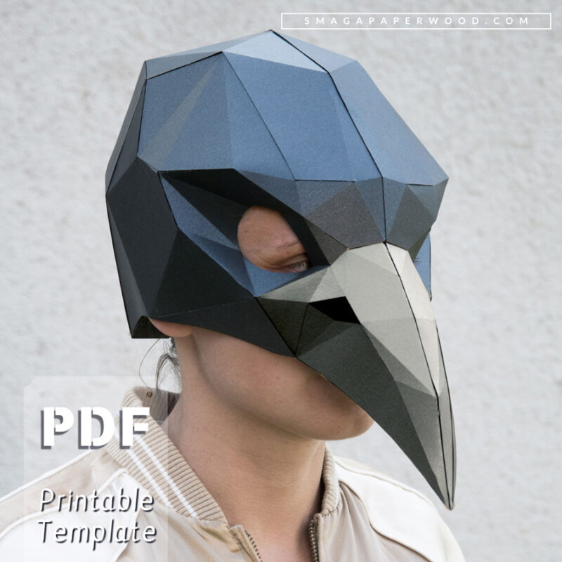 Geometric papercraft bird mask worn by a person against a neutral background. The mask, made from black and gray paper, features sharp angular facets and an elongated beak, resembling a raven or crow. Text on the image highlights that this is a printable PDF template for DIY crafting
