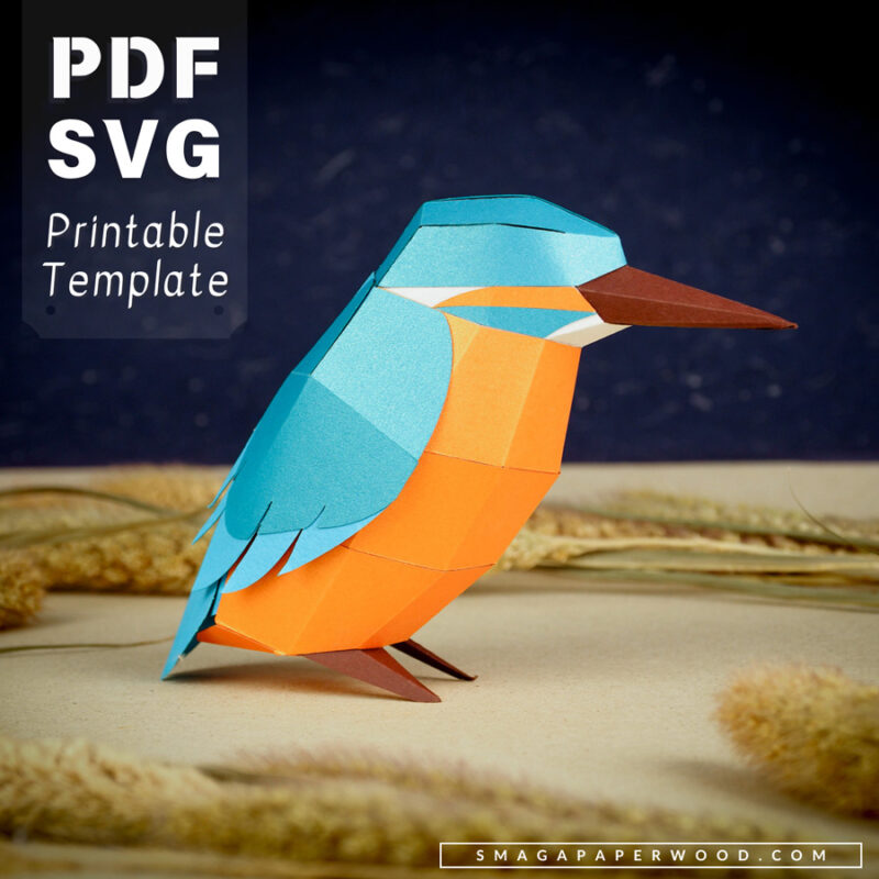 Geometric papercraft kingfisher bird standing on a neutral background. The bird is made of bright blue, orange and brown paper and has a faceted body and pointed beak. The image includes text highlighting that this is a printable PDF and SVG template,